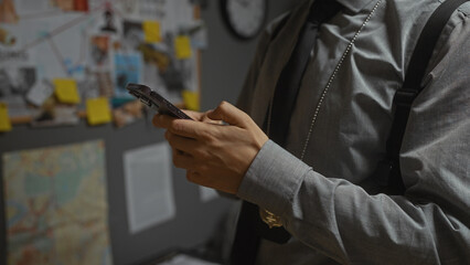 Man in detective's office analyzing evidence board with smartphone, evidencing investigation focus.