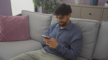 A smiling young man with a beard using a smartphone while seated on a gray couch indoors.