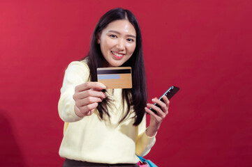 Fototapeta premium Asian woman holding a credit card and colorful shopping bags, looking to the side with a cheerful expression. Wearing a yellow top against a red background, suggesting a shopping or retail theme