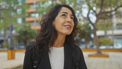 Mature hispanic woman smiling outdoors in a city park with trees and buildings in the background