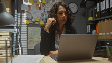 Focused woman detective working late on laptop in cluttered police station office with evidence...