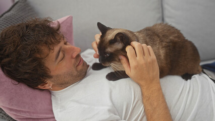 A young hispanic man lovingly pets his siamese cat while reclining on a sofa in a cozy living room.