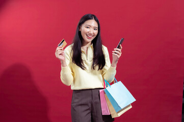 Asian woman smiling, holding a credit card and a smartphone, with colorful shopping bags on her...