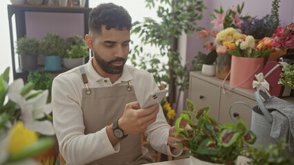 Hispanic man with a beard using a smartphone at a flower shop surrounded by various plants and colorful blooms.