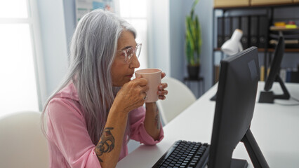 Elderly woman with grey hair in an office setting drinking from a mug while working on a computer