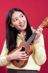 Fototapeta premium Asian woman playing a small ukulele with a joyful expression, dressed in a yellow sweater against a red background. Her posture and expression convey enjoyment and relaxation while making music.