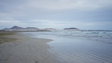 Blurry beach scene with mountains defocused in the background showcasing a calm seashore under a cloudy sky.