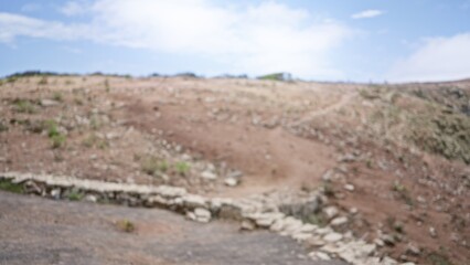 Blurred view of an arid countryside field with a defocused background, featuring rocks and scattered plants under a blue sky with white clouds.