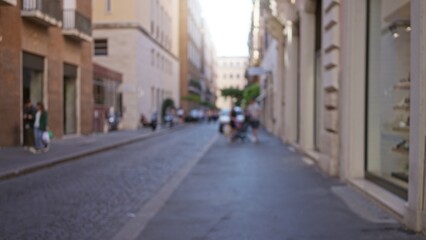 Obraz premium Blurred view of people walking on a cobblestone street in rome, italy, with historic buildings and storefronts creating a soft, defocused urban scene under bright daylight.