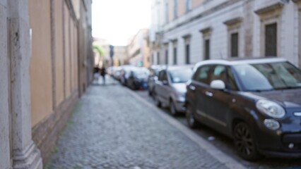 Blurred view of an old town street in rome, italy, showing defocused cars and pedestrians on a sunny day.