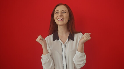 Cheerful young adult woman with red hair is celebrating over a solid red background, evoking joy...