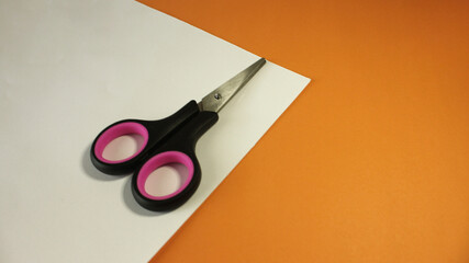 Black scissors with pink accents placed on a white sheet of paper, with a vibrant orange background.
