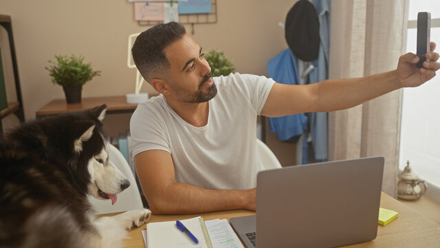 Hispanic man takes selfie with husky dog while working in cozy apartment living room