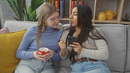 Two women sitting together on a couch in a cozy living room, one holding a red mug and the other using a smartphone.