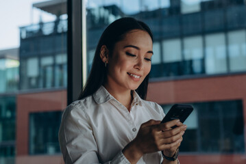 Portrait of satisfied businesswoman using smartphone near expansive window with city view