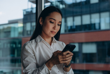 Asian female boss standing by modern office window and using smartphone in communicates with employee 