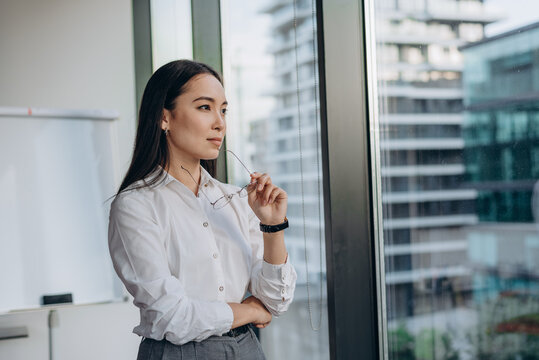 Young businesswoman stands by office window gazing thoughtfully at city skyline