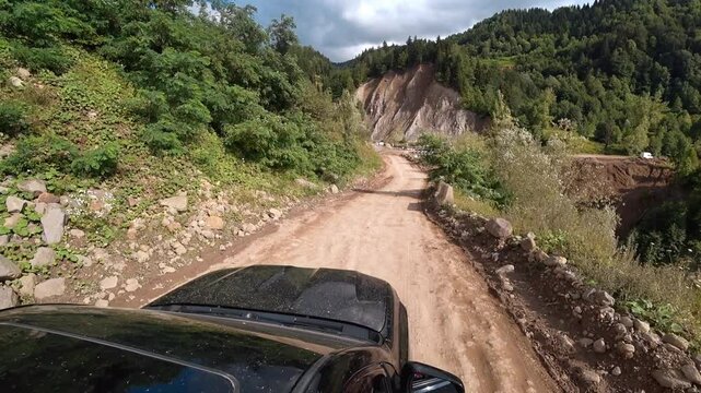 Black car driving on muddy road in mountains. Road is bumpy, and there are stones on side. Driver carefully navigates in difficult terrain. View of camera from roof of car.
