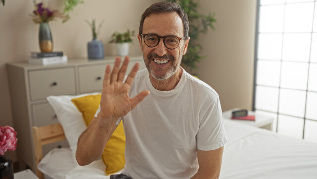 Middle-aged hispanic man waving in a bright bedroom with plants and books on a dresser beside the bed