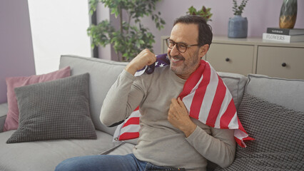 Middle-aged hispanic man with glasses smiles, draped in an american flag, sitting on a sofa in a...
