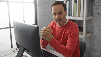 Middle-aged hispanic man in red sweater focused at his desk in an office with bookshelves and computer, embodying concentration and professionalism in a workplace setting.