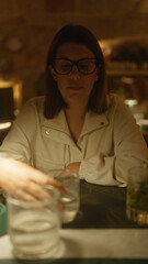 Young woman in a white jacket with glasses indoors at an italian restaurant, placing a glass on the table under warm lighting.
