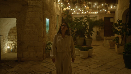 Naklejka premium A young hispanic woman walks through the beautifully lit streets of polignano a mare, puglia, italy at night, surrounded by plants and charming ancient stone buildings.