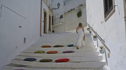 A young hispanic woman in a white dress climbs whitewashed stone steps adorned with colorful cushions in the charming old town of ostuni, puglia, italy, europe.