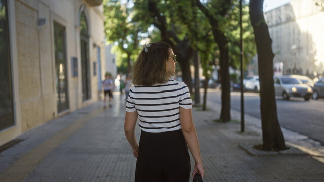 Fototapeta A young, beautiful hispanic woman walks down a tree-lined street in the charming town of lecce, puglia, italy on a sunny day.