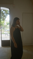 Young woman in a typical italian masseria house indoors, standing thoughtfully by a large open doorway with lush garden view.