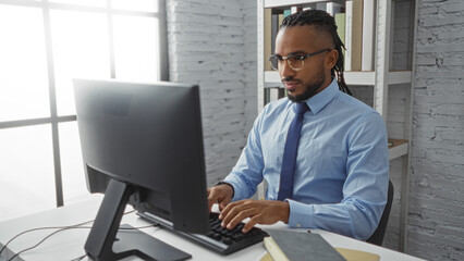 Obraz premium Young man working on computer in modern office wearing blue shirt and glasses focused on screen daytime setting with shelves in background suggesting professional environment