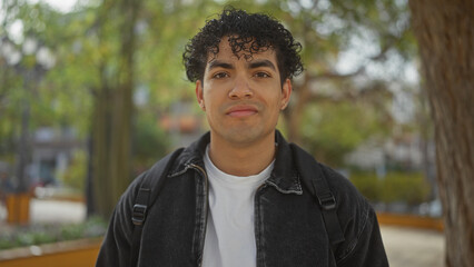 Handsome young hispanic man with curly hair in an urban park wearing a jacket and backpack