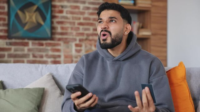 Portrait of emotional person in grey hoodie complaining about problems of using banking application. Concerned man making mistake while dealing password and entering account in electronic wallet.