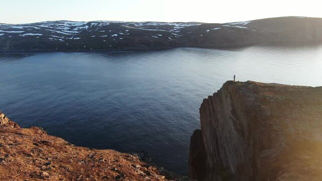 Aerial view of a serene lake with a man hiking along a rugged cliff, Skarsvag, Norway.