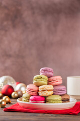 Multicolored macaroons in a plate with Christmas balls on a red napkin and cup of coffee. Christmas background. Vertical orientation