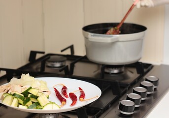 Close up to a plate with red peppers, zucchini and mushrooms in the kitchen, in the background something cooks in a pot