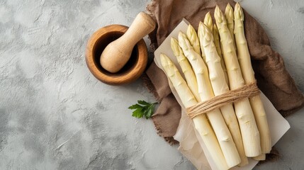 Freshly harvested organic white asparagus alongside a wooden mortar and paper on a light grey stone...