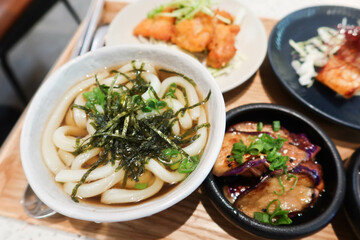 close up of soup udon with seaweed, mushroom appetiser, grilled salmon fish, eggplants and fried chicken in small plates bowls in a wooden tray