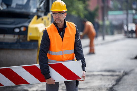 Construction worker in reflective safety gear holding a barrier while overseeing road work in a busy urban environment