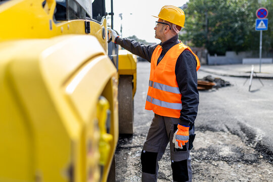 Construction worker inspecting heavy machinery on a road repair site during daylight in an urban area