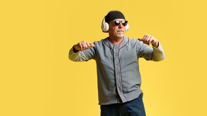 Elderly bearded man in cap and sunglasses listening to music in headphones and dancing against yellow studio background. Concept of human emotions, elderly lifestyle. Copy space, ad