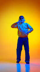 Full-length portrait of stylish elderly bearded man, in sunglasses and cap, listening to music in headphones and dancing against yellow background in neon. Concept of emotions, elderly lifestyle.