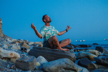 A man is peacefully sitting on a large rock at the beach, taking time to meditate and connect with his surroundings and nature