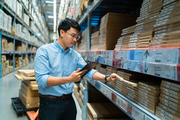 A man wearing a blue shirt is standing in a warehouse, looking at a shelf full of boxes. He is holding a tablet in his hand, possibly checking inventory or looking up information about the boxes