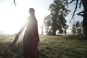 Fashion portarait of a young blonde girl in red strange dress. Autumn nature and tree branches. Sunrise and fog