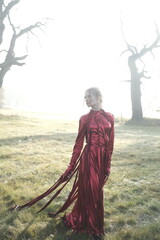 Fashion portarait of a young blonde girl in red strange dress. Autumn nature and tree branches. Sunrise and fog