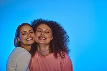 Two happy women embrace joyfully, sharing their love against a bright blue background in a studio.