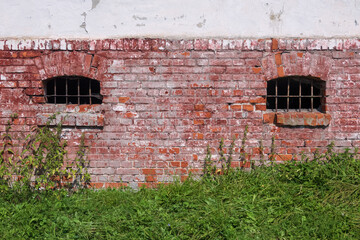 A fragment of the basement wall of an old brick house. There are two windows with bars and grass on the ground in front of the building. Background.