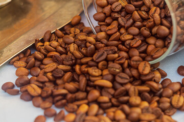 A pile of red roasted coffee beans spilled out of an overturned transparent glass jar onto the white surface of the tablecloth on table. One of the most important export products in some countries