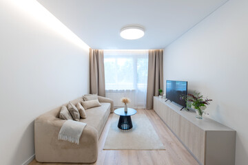 Modern Home Living Room Interior with Open TV Area and TV on the Wooden Desk, Sofa, And Coffee Table. Flowers on the Table. Windows and Curtains in Background. Hardwood Floor, Carpet on the ground